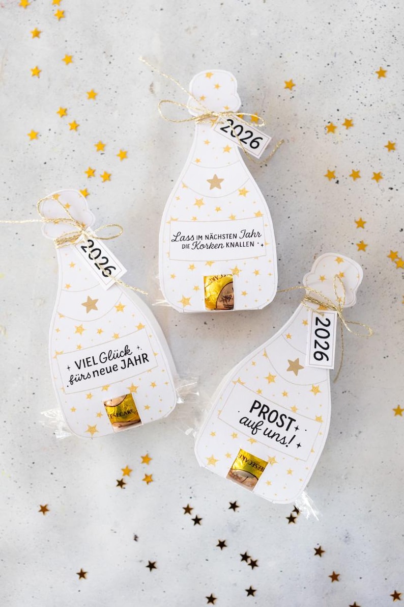 Puede incluir: Tres etiquetas de regalo con forma de botella de champ&aacute;n con patrones de estrellas doradas y el a&ntilde;o "2026" en las etiquetas. Cada etiqueta tiene una frase alemana diferente, como "&iexcl;Feliz A&ntilde;o Nuevo!"
