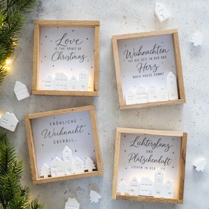 Puede incluir: Cuatro cajas decorativas cuadradas de madera con siluetas de casas de cerámica blanca y fondos iluminados. Cada caja muestra texto en alemán o inglés, incluyendo Love is the spirit of Christmas, Weihnachten ist die Zeit, in der das Herz nach Hause kommt, Fröhliche Weihnacht überall y Lichterglanz und Plätzchenduft.
