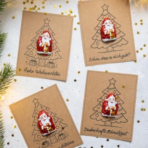 Puede incluir: Cuatro tarjetas rectangulares de papel kraft con dibujos de líneas negras de árboles de Navidad y regalos. Cada tarjeta tiene un Papá Noel de chocolate envuelto en papel de aluminio. Las tarjetas incluyen texto en alemán: frohe Weihnachten, Schön, dass es dich gibt, Ein Gruß vom Nikolaus y Zauberhafte Adventszeit.