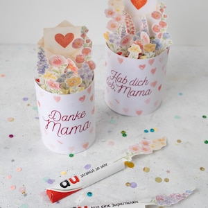 Könnte beinhalten: Zwei weiße Papierbehälter mit Blumenarrangements und Herzakzenten. Einer sagt "Danke Mama" und der andere "Hab dich lieb, Mama". Zwei Schokoriegel mit Blumendekorationen sind ebenfalls vorhanden.