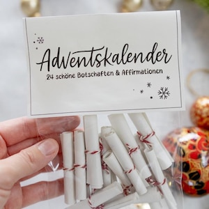Può includere: Un sacchetto di plastica trasparente pieno di rotoli di carta bianca legati con spago rosso. Il sacchetto ha un'etichetta bianca con il testo "Adventskalender" e "24 schöne Botschaften & Affirmationen". Decorazioni natalizie sullo sfondo.