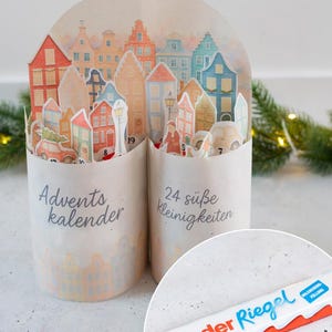 Könnte beinhalten: Adventskalender mit einem Stadtbild-Design, mit bunten Häusern und dem Text "Advents kalender" und "24 süße Kleinigkeiten". Eine Kinder Riegel Schokoriegel ist ebenfalls zu sehen.