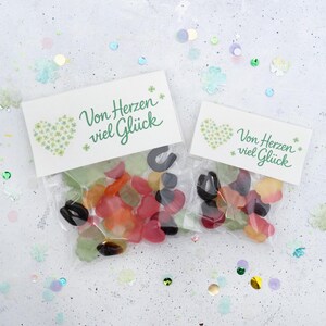 May include: Two clear cellophane bags filled with colorful gummy candies, each topped with a white card. The cards read "Von Herzen viel Glück" in green script, with a heart and shamrock design. Confetti surrounds the bags.