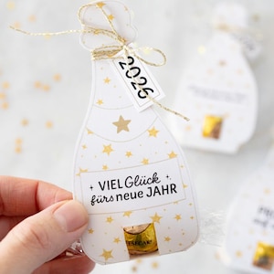 Puede incluir: Una etiqueta de regalo blanca con forma de botella de champ&aacute;n con detalles de estrellas doradas y el a&ntilde;o "2026" impreso en una etiqueta peque&ntilde;a. La etiqueta tambi&eacute;n tiene el texto "VIEL Gl&uuml;ck f&uuml;rs neue JAHR". Una cinta dorada est&aacute; atada en la parte superior.