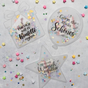 以下が含まれることがあります： コンフェッティとテキストが詰められた3つの透明な装飾的な形：ギフトボックス、風船、星。ギフトボックスには「FÜR DICH soll es Konfetti regnen」と書かれています。風船には「Gönn dir etwas Schönes」と書かれています。星には「EIN KLEINER Wunsch-erfüller」と書かれています。
