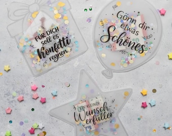 Op de afbeelding: Drie heldere, decoratieve vormen gevuld met confetti en tekst: een geschenkdoos, een ballon en een ster. De geschenkdoos zegt "FÜR DICH soll es Konfetti regnen". De ballon zegt "Gönn dir etwas Schönes". De ster zegt "EIN KLEINER Wunsch-erfüller".