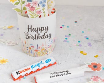 Op de afbeelding: Een witte papieren beker met de tekst "Happy Birthday" en bloemenillustraties, met papieren bloemenuitsparingen. Twee Kinder Riegel chocoladerepen en twee berichtenstokjes met bloemaccenten zijn ook aanwezig.