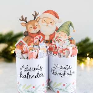 Könnte beinhalten: Zwei weiße zylindrische Behälter mit der Aufschrift Advents kalender und 24 süße Kleinigkeiten. Die Behälter sind mit verschiedenen weihnachtlichen Papieraufstellern gefüllt, darunter der Weihnachtsmann, ein Rentier, ein Elf und ein Schneemann.