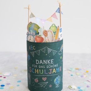 May include: A cylindrical green container with school-themed illustrations and the German text "DANKE F&Uuml;R DAS SCH&Ouml;NE SCHULJAHR." The container is decorated with a globe, apple, pencil, and a rainbow. Small flags are attached to the top.
