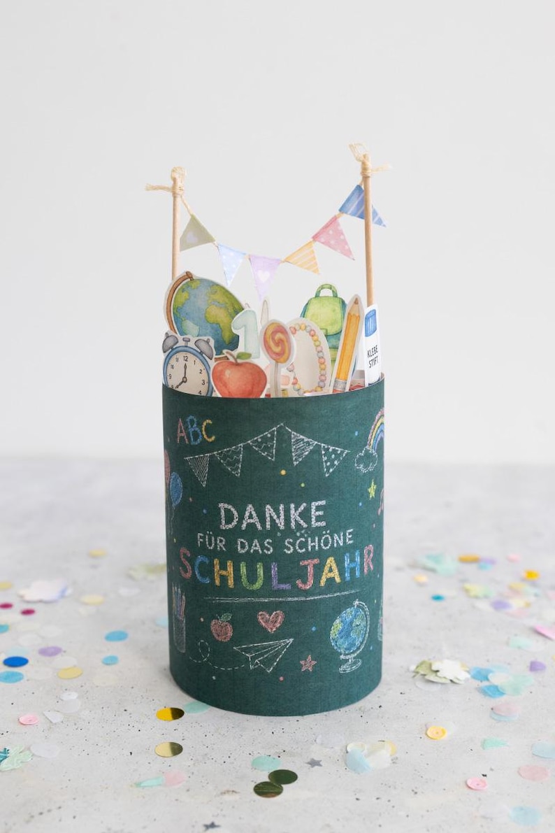 May include: A cylindrical green container with school-themed illustrations and the German text "DANKE F&Uuml;R DAS SCH&Ouml;NE SCHULJAHR." The container is decorated with a globe, apple, pencil, and a rainbow. Small flags are attached to the top.