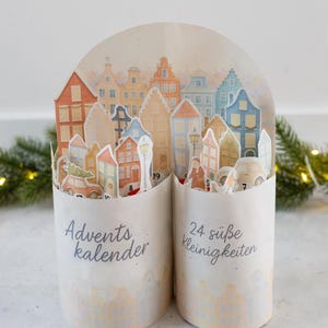 Könnte beinhalten: Adventskalender mit Stadtbild-Design. Der Papierkalender zeigt eine Reihe farbenfroher Häuser und Gebäude. Der Text "Advents kalender" und "24 süße Kleinigkeiten" ist auf der Vorderseite aufgedruckt.