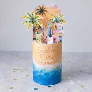 May include: A cylindrical card featuring a beach scene with palm trees, a surfboard, a cocktail, and a signpost. The base shows a sandy beach transitioning into blue ocean waves. The text Endlich Ruhe (stand) is printed on the front of the cylinder.