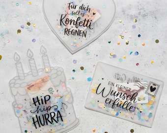 Op de afbeelding: Drie doorschijnende acryl cadeauhouders met kleurrijke confetti erin. Eén is hartvormig met de tekst "Für dich soll es Konfetti REGNEN", één is een taart met drie kaarsjes en "Hip hip HURRA", en één is een envelop met "EIN KLEINER Wunscherfüller".