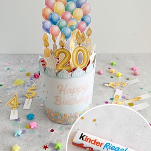 May include: A birthday-themed pop-up centerpiece with colourful balloons, candles, and the number 20. The words "Happy Birthday" are displayed on the front. A Kinder Riegel chocolate bar is also present.