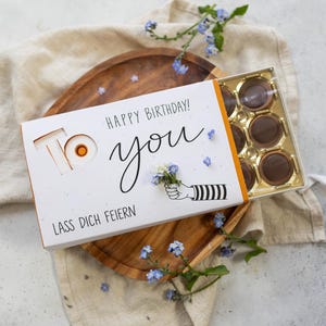 Può includere: Una scatola regalo bianca con le scritte "Happy Birthday! To you" e "Lass Dich Feiern" stampate sopra. La scatola è aperta, rivelando cioccolatini. La scatola è su un vassoio di legno, decorato con fiori blu.