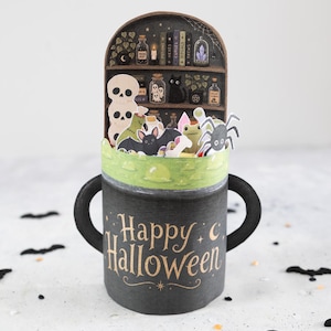 Könnte beinhalten: Eine dreidimensionale Papierbastelkarte in Form eines schwarzen Kessels mit zwei Griffen. Auf der Vorderseite steht der goldene Schriftzug Happy Halloween. Oben befindet sich ein Pop-up-Regal mit Miniatur-Zaubertrankflaschen, Totenköpfen, einer schwarzen Katze und einer Spinne.