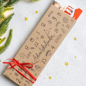 Könnte beinhalten: Ein rechteckiger Adventskalender aus braunem Papier mit einer roten Schleife am unteren Ende. Die Oberfläche zeigt schwarze Zahlen, weihnachtliche Symbole und den Text Adventskalender. Ein Teil eines Kinder-Schokoriegels ist zu sehen.