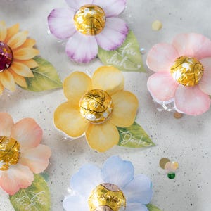 Peut inclure: Une collection de fleurs en papier aux couleurs pastel variées, chacune dotée d'une truffe en chocolat Lindt Lindor emballée dans du papier doré au centre. Des feuilles en papier vert sont attachées, certaines avec le texte Danke ou für dich.
