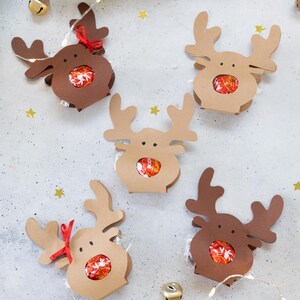 Puede incluir: Una colecci&oacute;n de cajas de regalo con forma de reno hechas de cartulina marr&oacute;n y beige. Cada caja contiene caramelos envueltos y est&aacute; decorada con una cinta roja. Las cajas est&aacute;n dispuestas sobre una superficie gris claro con decoraciones festivas.