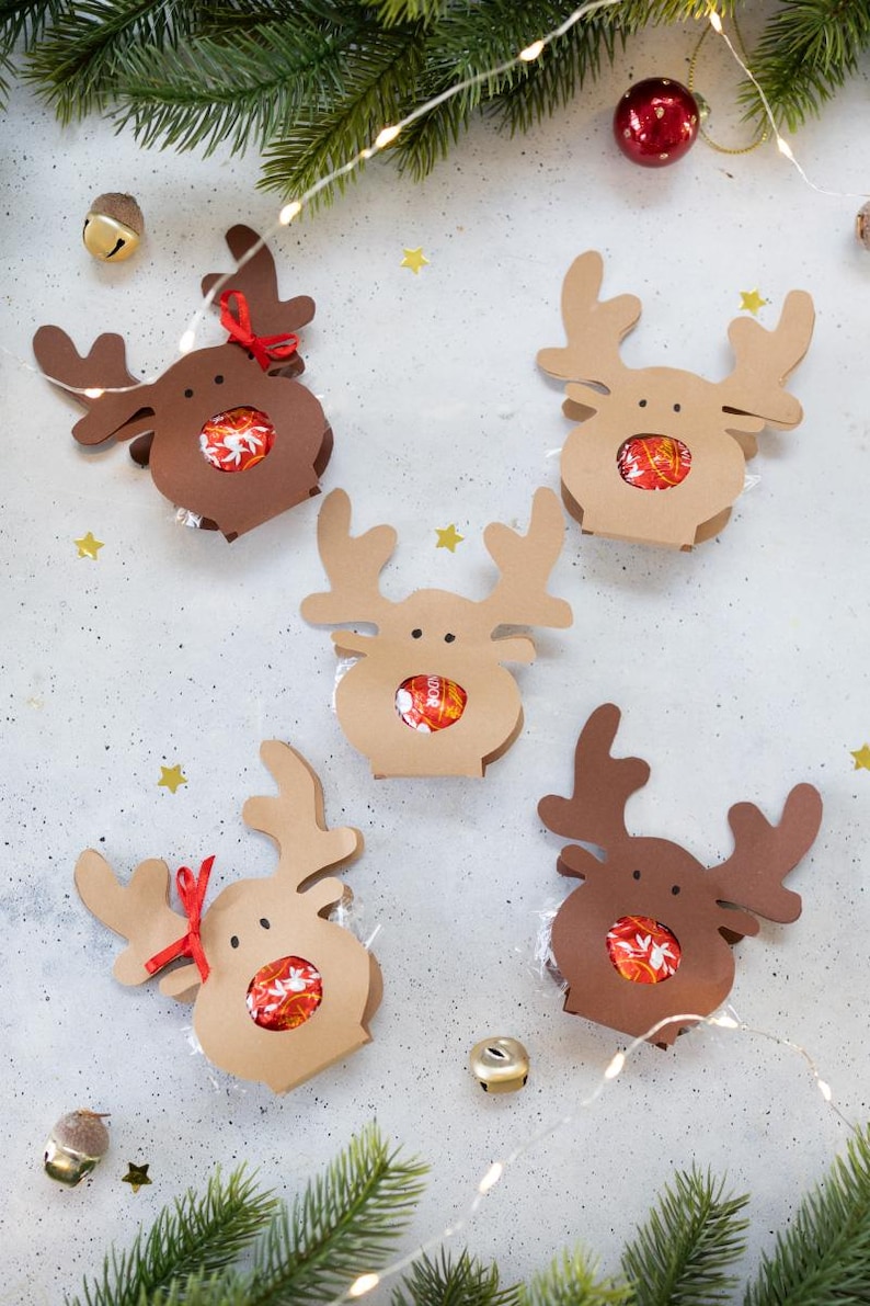 Puede incluir: Una colecci&oacute;n de cajas de regalo con forma de reno hechas de cartulina marr&oacute;n y beige. Cada caja contiene caramelos envueltos y est&aacute; decorada con una cinta roja. Las cajas est&aacute;n dispuestas sobre una superficie gris claro con decoraciones festivas.