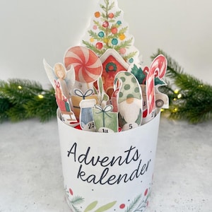 Könnte beinhalten: Ein weißer Adventskalender mit Aquarellillustrationen von Weihnachtselementen. Der Kalender zeigt einen Weihnachtsbaum, einen Engel, einen Zwerg, Geschenke und Zuckerstangen. Die Worte "Advents kalender" sind aufgedruckt.
