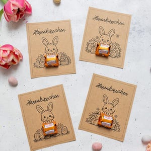 Könnte beinhalten: Vier Karten aus Kraftpapier mit einer handgezeichneten Osterhasen-Illustration und dem Schriftzug "Hasenküsschen". Jede Karte enthält ein verpacktes Schokoladenbonbon. Rosa Tulpen und pastellfarbene Ostereier sind um die Karten herum angeordnet.