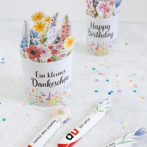Puede incluir: Vasos de papel blancos con diseños florales y las frases "Happy Birthday" y "Ein kleines Dankeschön". Las tazas están llenas de flores de papel. También hay tres barras de chocolate con acentos florales y el texto "du".