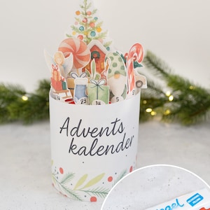 Peut inclure: Un calendrier de l'Avent blanc avec un sapin de Noël pop-up, des cadeaux et des cannes en bonbon. Le calendrier porte l'inscription "Advents kalender" et est orné d'illustrations festives. Une barre chocolatée Kinder Riegel est dans le coin.
