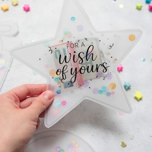 Könnte beinhalten: Ein klarer, sternförmiger Geschenkkartenhalter mit dem Text "For a wish of yours". Der Halter enthält bunte Konfetti und einen kleinen Geldbetrag. Bunte sternförmige Konfetti sind verstreut.