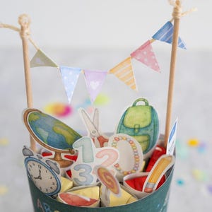 May include: A decorative paper cup filled with school-themed cutouts, including a globe, backpack, scissors, and numbers. A small banner with pastel flags tops the cup. The cup has the text "ABC" and "DANKE F&Uuml;R DAS SCH&Ouml;NE".