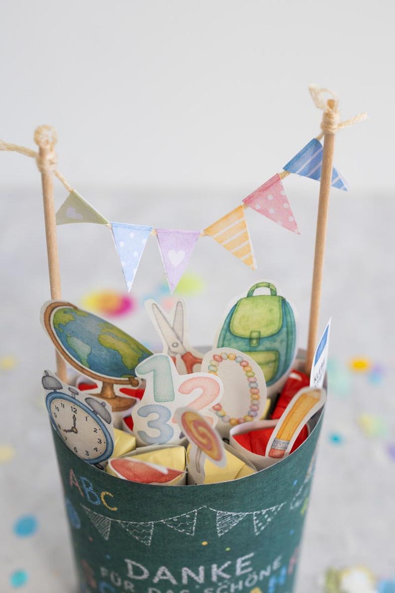May include: A decorative paper cup filled with school-themed cutouts, including a globe, backpack, scissors, and numbers. A small banner with pastel flags tops the cup. The cup has the text "ABC" and "DANKE F&Uuml;R DAS SCH&Ouml;NE".