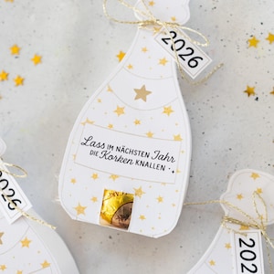 Puede incluir: Etiquetas de regalo blancas en forma de botella de champ&aacute;n con patrones de estrellas doradas y el a&ntilde;o "2026" en una etiqueta. Las etiquetas tienen una cuerda dorada y un peque&ntilde;o caramelo de chocolate en el interior. El texto "Lass im n&auml;chsten Jahr die Korken knallen" est&aacute; impreso.