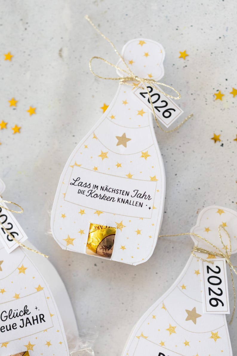 Puede incluir: Etiquetas de regalo blancas en forma de botella de champ&aacute;n con patrones de estrellas doradas y el a&ntilde;o "2026" en una etiqueta. Las etiquetas tienen una cuerda dorada y un peque&ntilde;o caramelo de chocolate en el interior. El texto "Lass im n&auml;chsten Jahr die Korken knallen" est&aacute; impreso.