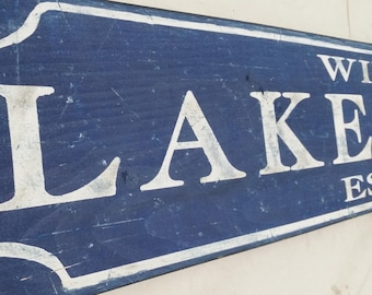 Op de afbeelding: Een rechthoekig houten bord met een verweerde blauwe achtergrond en witte letters. Het bord leest "LAKE" in grote letters, met "WILD" en "EST" erboven en eronder. Het bord heeft een witte rand.