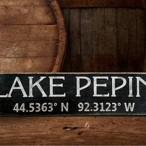 May include: Black wooden sign with white text that reads "LAKE PEPIN 44.5363° N 92.3123° W". The sign is sitting on a wooden surface with two wooden barrels in the background.