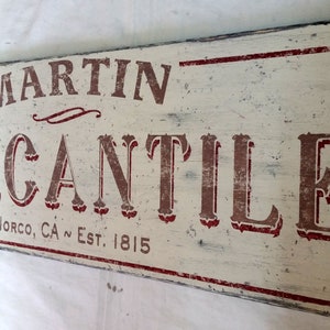 Op de afbeelding: Een wit houten bord met rode letters waarop "Martin Mercantile" en "Norco, CA Est. 1815" staat.