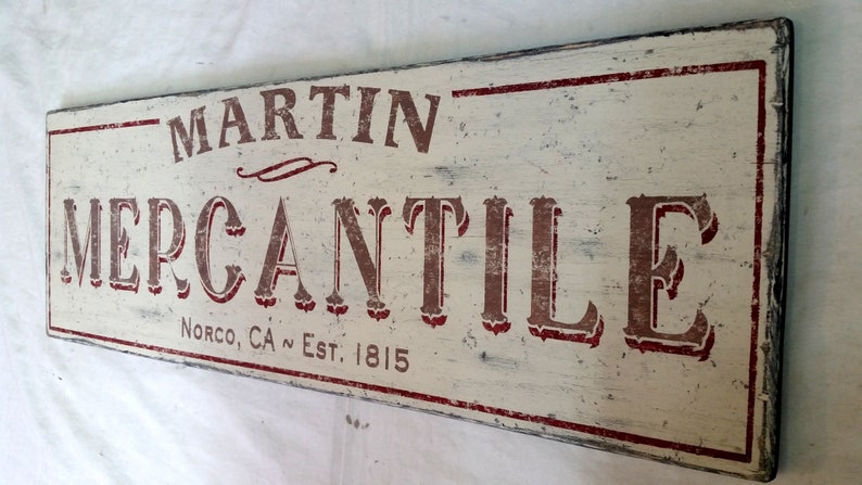 Op de afbeelding: Een wit houten bord met rode letters waarop "Martin Mercantile" en "Norco, CA Est. 1815" staat.