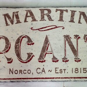 Op de afbeelding: Een wit houten bord met rode en bruine letters met de tekst "MARTIN MERCANTILE" en daaronder de tekst "NORCO, CA ~ EST. 1815".