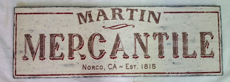 Op de afbeelding: Een wit houten bord met rode en bruine letters met de tekst "MARTIN MERCANTILE" en daaronder de tekst "NORCO, CA ~ EST. 1815".