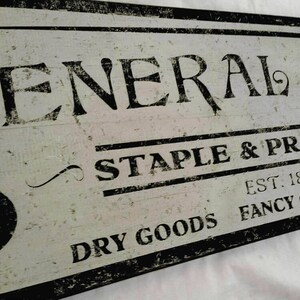 Puede incluir: Un letrero de estilo vintage para una tienda general, pintado en blanco y negro con el texto "GENERAL STORE", "STAPLE & PROVISIONS", "EST. 1894", "DRY GOODS FANCY GROCERY SPICES", y un s&iacute;mbolo de 5&cent;.