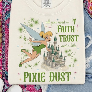 Könnte beinhalten: Ein cremefarbenes T-Shirt mit Rundhalsausschnitt und einer Grafik einer Fee mit blonden Haaren und grüner Kleidung, die neben einem Schloss fliegt. Der Text lautet: all you need is FAITH TRUST and a little PIXIE DUST. Das Shirt hat ein Comfort Colors Etikett.