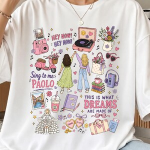 May include: A white t-shirt featuring a colorful graphic print of two figures with raised arms, surrounded by illustrations of a pink scooter, a record player, a camera, a bouquet, books, a suitcase, and text including "HEY NOW! HEY NOW!" and "Sing to me PAOLO".
