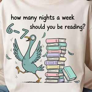 Könnte beinhalten: Ein cremefarbenes T-Shirt mit einer Cartoon-Illustration eines blauen Vogels, der auf einen Stapel Bücher zuläuft. Der Text lautet: „Wie viele Nächte pro Woche solltest du lesen?“ mit den Zahlen „6-7“ über dem Vogel.