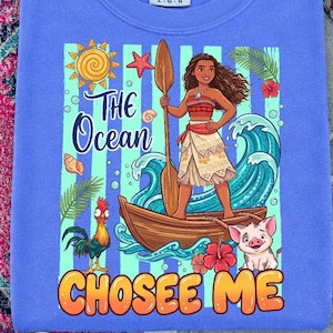 Könnte beinhalten: Blaues Langarmshirt mit einem Aufdruck, der eine Figur in einem Boot, Wellen und den Text "THE Ocean CHOSEE ME" zeigt. Das Design umfasst eine Sonne, einen Seestern und tropische Blumen. Marke Comfort Colors.