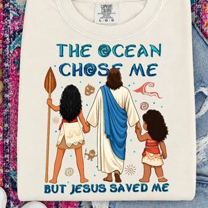 May include: Cream-colored Comfort Colors sweatshirt with the text "THE OCEAN CHOSE ME BUT JESUS SAVED ME" in blue. The graphic depicts three figures, including Jesus, and two girls. The sweatshirt is a crew neck and has long sleeves.