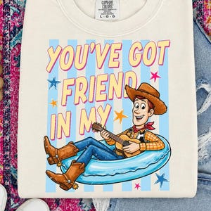 May include: A cream-colored crewneck t-shirt featuring a graphic of a cowboy character playing a guitar while sitting in a blue inner tube. The text reads YOU'VE GOT FRIEND IN MY above the character, surrounded by small blue and pink stars.