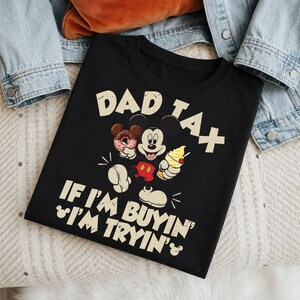 May include: Black t-shirt with the text "DAD TAX" and "IF I'M BUYIN' I'M TRYIN'" in a playful font. The shirt features a graphic of Mickey Mouse with a donut, ice cream, and his signature gloves and shorts. A denim jacket is in the background.