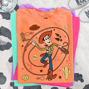 May include: An orange t-shirt featuring a graphic of the character Woody from Toy Story, who is holding a lasso. The design includes a sheriff badge, a cowboy hat, a boot, a cactus, a horseshoe, and the name Woody written in cursive script.