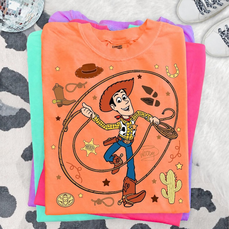 May include: An orange t-shirt featuring a graphic of the character Woody from Toy Story, who is holding a lasso. The design includes a sheriff badge, a cowboy hat, a boot, a cactus, a horseshoe, and the name Woody written in cursive script.