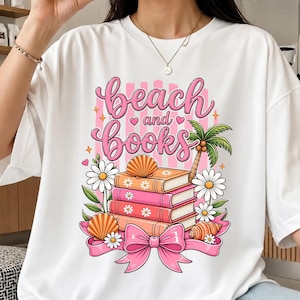 May include: White t-shirt with a colourful graphic design. The design features the words "beach and books" in pink script, a stack of books, seashells, daisies, and a palm tree. A pink bow is at the bottom.